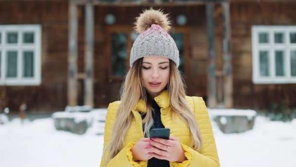 Retrato de una mujer joven utilizando la aplicación en el teléfono inteligente, sonriendo y mensajes de texto en el teléfono móvil. Mujer vestida con un abrigo de invierno cerca de una casa de campo mirando a la cámara. Comunicación, Viajes, Concepto de Estilos de Vida — Vídeo de stock