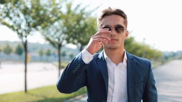 Joven empresario exitoso en gafas de sol caminando por la ciudad. Se quita las gafas y mira a la cámara. Elegante atuendo elegante. Ser un jefe, estilo de vida exitoso — Vídeos de Stock