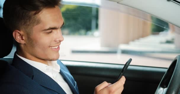 Jonge knappe zakenman gebruik mobiele telefoon in de auto. Gelukkig jonge man slaagt en schrijft een bericht. Het concept van communicatie, succesvol bedrijf, mobiele technologie — Stockvideo
