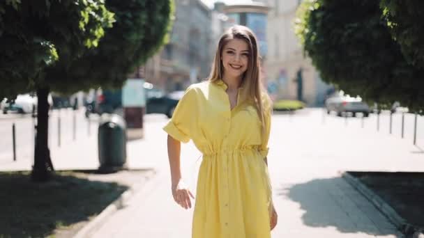 Mooi meisje in gele jurk wandelen langs de straat straat stad. Onscherpe achtergrond zonnige dag zomer zonneschijn vrouw die lacht vrij mooie blik op zoek camera glimlacht blij gezicht van emotionele ontspanning — Stockvideo