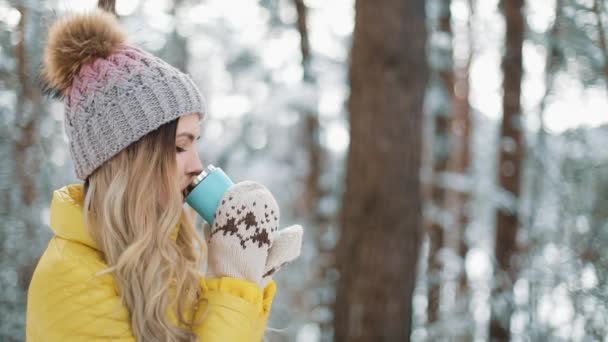 Mulher bonita no chapéu de inverno gosta da neve e bebe bebida quente de pé do lado de fora na neve na floresta. Menina desfrutando de inverno ao ar livre. Férias de Natal — Vídeo de Stock