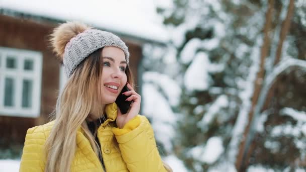 Donna sorridente parla al telefono in piedi fuori sul cortile sotto la neve che cade. Donna che indossa un cappotto invernale vicino a una casa di campagna. Comunicazione, Viaggi, Stili di Vita Concetto. Girato su Red Epic — Video Stock