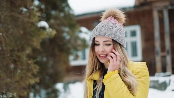 Mulher sorridente fala ao telefone do lado de fora no quintal sob a neve caindo. Mulher vestindo um casaco de inverno perto de uma casa de campo. Comunicação, Viagens, Estilo de Vida Conceito. Tiro no épico vermelho — Vídeo de Stock