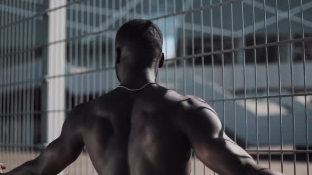 Соблазнительный афроамериканец с мускулами позирует с обнаженной грудью перед забором. Motivation, pumping body, sports, gym, success, street workout, muscles — стоковое видео
