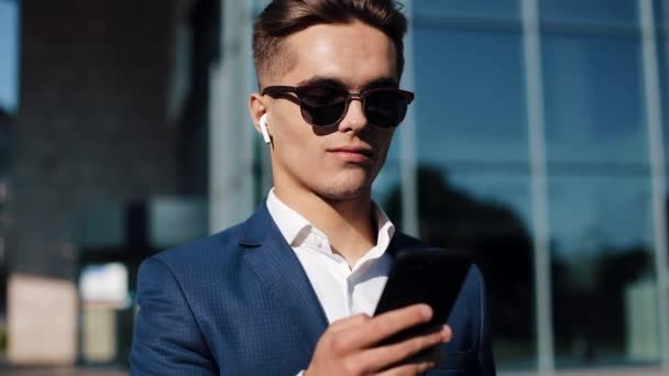 Jeune homme d'affaires en lunettes de soleil utilisant un smartphone et marchant dans la rue. Une vie réussie. Style d'affaires, voyageur, communication, style de vie moderne. Mode de vie actif . — Video