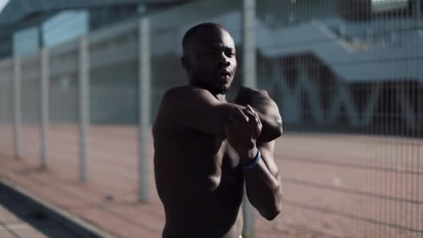 L'atleta afroamericano fa ginnastica mattutina all'aperto vicino al sole al rallentatore. Allenamento in piedi sulla strada vicino allo stadio sportivo. Stile di vita sano forza concetto motivazione — Video Stock