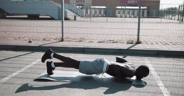 Уличная тренировка. Атлетичный афроамериканец делает отжимания и упражнения на свежем воздухе. Черный мужчина занимается спортом возле стадиона. Тренировки, фитнес, бег, мотивация — стоковое видео