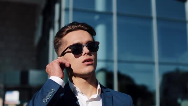 Jeune homme d'affaires attrayant parlant au téléphone en utilisant des écouteurs sans fil marchant à l'extérieur. Un homme heureux parle. Communication, finance, hommes d'affaires, stagiaires, PDG. Ferme là. Tourné sur Red Epic — Video