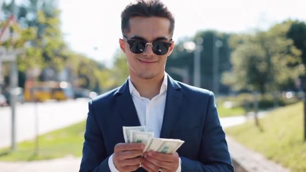 Joven caballero feliz en gafas de sol y un traje contando dinero y caminando por la calle. Selebrando su exitosa victoria con un montón de dólares — Vídeo de stock