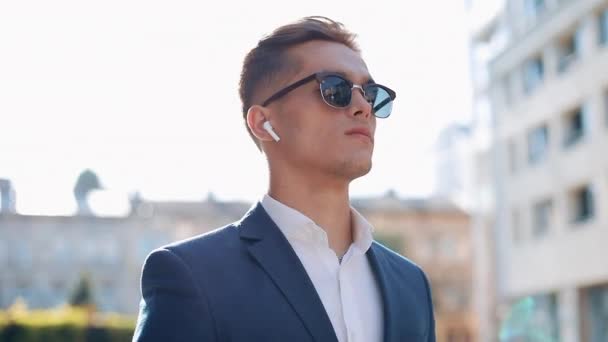 Молодой бизнесмен в солнечных очках слушает музыку на своем смартфоне на улице. Он идет рядом с офисным зданием. Коммуникации, аудиокниги, музыка, бизнесмены. Shot on Red Epic — стоковое видео