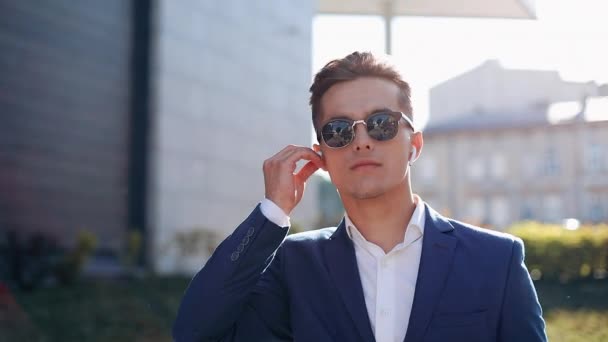 Feliz empresario con gafas de sol escuchando la música en su smartphone al aire libre. Camina cerca del edificio de oficinas. Comunicación, audiolibros, música, gente de negocios. Disparo en la épica roja — Vídeo de stock