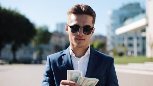 Junger, fröhlicher Geschäftsmann mit Sonnenbrille und Anzug, der Geld zählt und auf der Straße spazieren geht. er selebriert seinen erfolgreichen Sieg mit einer Menge Dollar — Stockvideo