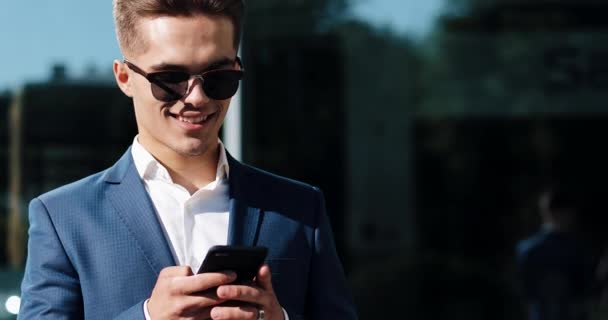 Homme utilisant l'application d'affaires sur téléphone intelligent debout à l'extérieur. Beau jeune homme d'affaires communiquant sur smartphone confiant souriant.Tourné sur Red Epic — Video