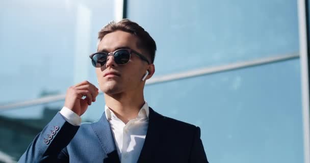 Atraktivní mladý podnikatel pomocí bezdrátové sluchátka stojí venku. Nosí brýle a vypadá pryč. Komunikace, finance, podnikatelé, stážistů, generální ředitel. Zblízka. Střílel na Red Epic — Stock video