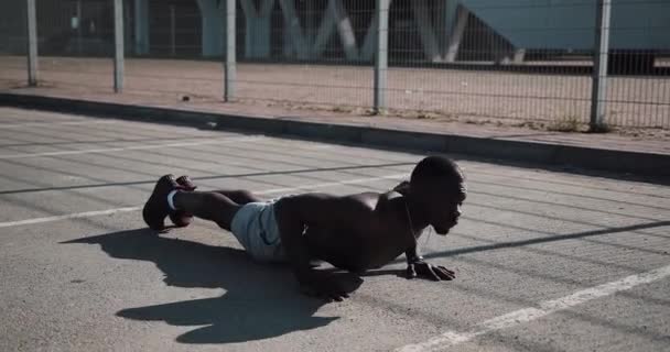 Уличная тренировка. Атлетичный афроамериканец делает отжимания и упражнения на свежем воздухе. Черный мужчина занимается спортом возле стадиона. Тренировки, фитнес, бег, мотивация — стоковое видео