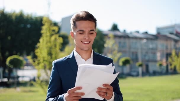 Joven hombre de negocios feliz en un traje elegante examina cuidadosamente los papeles al aire libre. Estilo de vida exitoso, contrato, responsabilidad. Negocios, personas, concepción del papeleo — Vídeo de stock