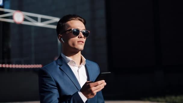Genç businesman smartphone kullanarak ve sokakta yürürken güneş gözlüğü. Başarılı bir hayat. İş tarzı, Gezgin, iletişim, modern yaşam tarzı. Aktif yaşam tarzı — Stok video