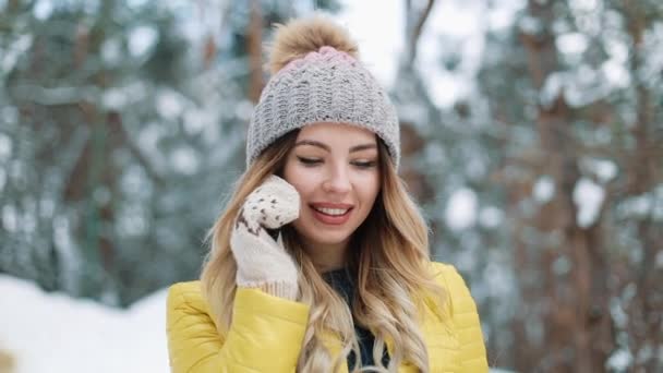 Улыбающаяся девушка в зимней шляпе разговаривает по смартфону, гуляя по лесу, покрытому снегом. Молодая красивая женщина разговаривает по мобильному телефону, гуляя в зимнем парке — стоковое видео