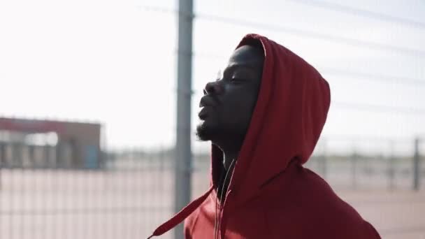 Man loopt bij zonsondergang in de stad. African American mannelijke atleet loper het dragen van de Rode hoodie. Uitoefening buitenshuis fitness tracker wearable technologie — Stockvideo