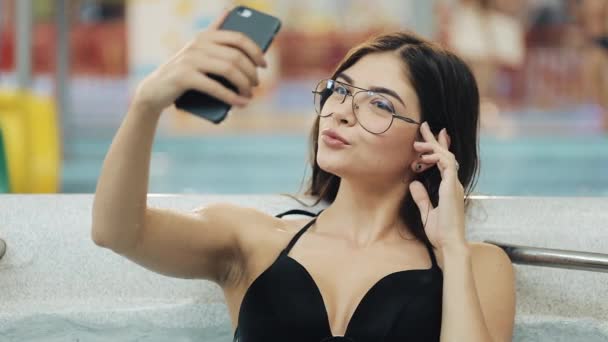 Schöne junge Frau in Schwarz macht Selfie auf ihrem Smartphone in einem brandneuen Whirlpool oder Schwimmbad — Stockvideo