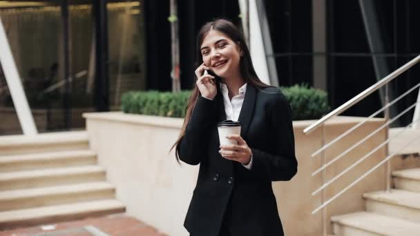 Веселая молодая деловая женщина, стоящая рядом с офисным центром, разговаривающая по смартфону. Она смотрит в камеру. Концепция: новый бизнес, коммуникация, банкир. Снаружи, медленная съемка — стоковое видео