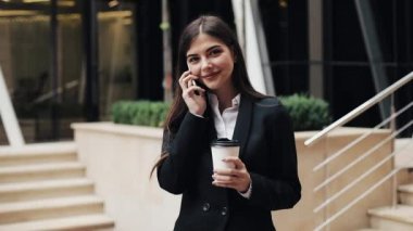 Office center yanında duran, belgili tanımlık smartphone üzerinde konuşurken neşeli genç iş kadını. Kameraya bakıyor. Konsept: yeni iş, iletişim, bankacı. Dış, ağır çekim
