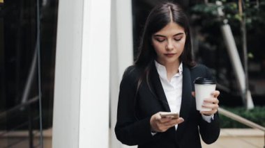Profesyonel genç iş kadını Smartphone ve içecek kahve kullanarak kentsel sokakta yürümek. Konsept: yeni iş, iletişim, bankacı. Dış, ağır çekim