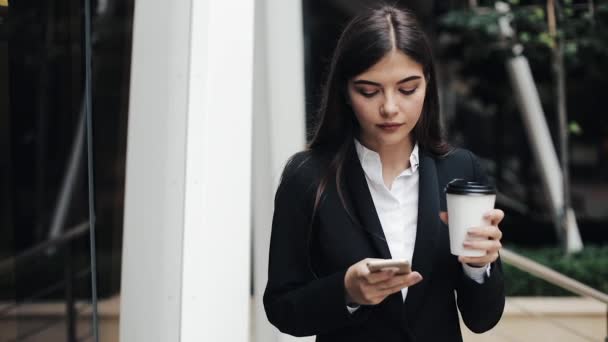 Професійна молода бізнес-леді, що йде по міській вулиці, використовує смартфон і п'є каву. Концепція: новий бізнес, комунікація, банкір. Зовні, повільний рух — стокове відео