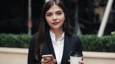 Profesyonel genç iş kadını Smartphone ve içecek kahve kullanarak kentsel sokakta yürümek. Kameraya bakıyor. Konsept: yeni iş, iletişim, bankacı. Dış, ağır çekim