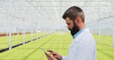 Erkek araştırmacı çalışmaları bitkiler sera içinde bir tablet konumu olan sakallı. O içine belgili tanımlık fotoğraf makinesi ve gülümsüyor seyir kameraya, döner