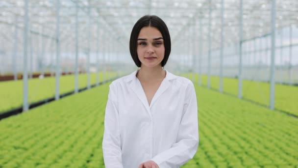 Jordbruksingenjören i växthus klädd i vit rock tittar in i kameran. Porträtt av en leende kvinna i växthus — Stockvideo