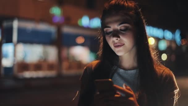 Wanita muda yang menarik menggunakan smartphone berdiri di jalan kota malam bereaksi bahagia dengan pesan itu. Teknologi modern, sukses — Stok Video