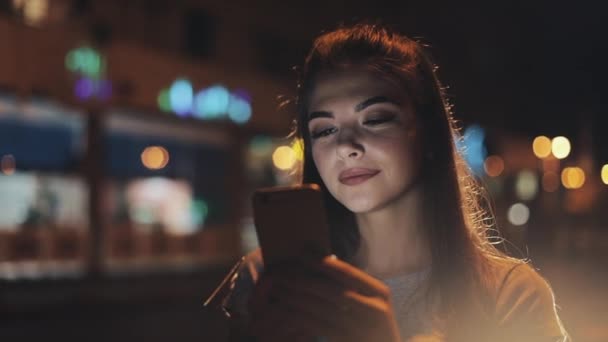 迷人的年轻女子使用智能手机站在夜间镇的街道上高兴地响应的信息。现代技术, 成功 — 图库视频影像