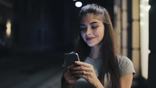 Приваблива молода жінка, використовуючи смартфон, що стоїть на вулиці нічного міста, з радістю реагує на повідомлення. Сучасні технології, успішні — стокове відео
