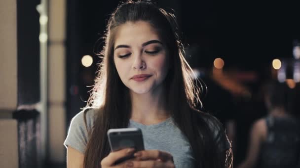 Jovem atraente usando smartphone em pé na rua da cidade noturna reagindo alegremente à mensagem. Tecnologias modernas, bem sucedidas — Vídeo de Stock