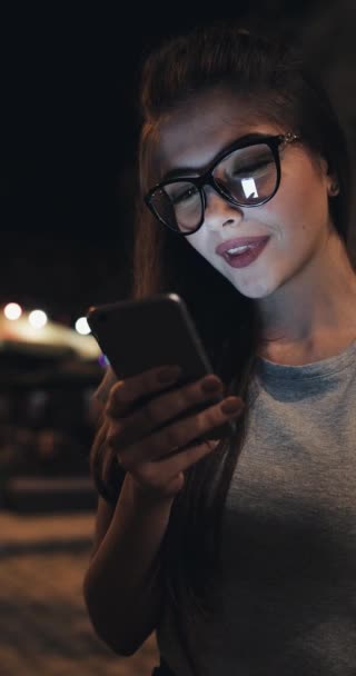 Attraente giovane donna in occhiali utilizzando smartphone seduto sulla panchina sulla strada della città di notte che reagisce felicemente al messaggio. Tecnologie moderne, di successo — Video Stock