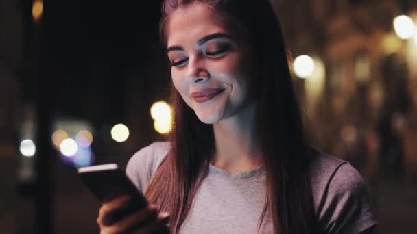 Jovem atraente usando smartphone em pé na rua da cidade noturna reagindo alegremente à mensagem. Tecnologias modernas, bem sucedidas — Vídeo de Stock