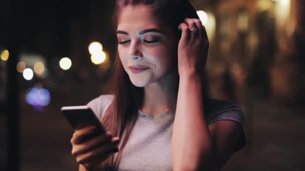 Jovem atraente usando smartphone e olhando para a câmera. Ela estava na rua da cidade noturna reagindo alegremente à mensagem. Tecnologias modernas, bem sucedidas — Vídeo de Stock