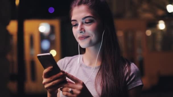 Mujer joven atractiva con auriculares en los oídos escuchando música y usando un teléfono inteligente. Noche borrosa — Vídeo de stock