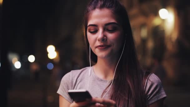Mujer joven atractiva con auriculares en los oídos escuchando música y usando un teléfono inteligente. Noche borrosa — Vídeo de stock