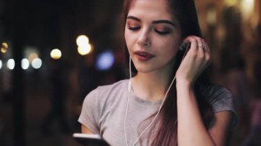Çekici genç kadın açık havada akşam akıllı telefon üzerinde müzik dinlemek. Gülümseyen kız dışında yeni şarkıları zevk geceleri ayakta portresi.