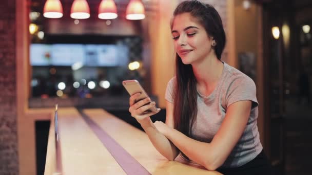 Jovem mulher sentada no bar usando seu telefone inteligente ao lado do sinal de barra de néon. Mulher desfrutando da vida noturna enquanto se comunica com amigos em seu smartphone — Vídeo de Stock