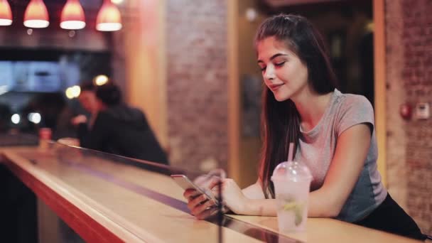 Mujer joven sentada en el bar usando su teléfono inteligente al lado del letrero del bar de neón. Mujer disfrutando de la vida nocturna mientras se comunica con amigos en su teléfono inteligente — Vídeos de Stock