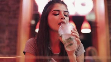 Genç kadın otururken bar, lemanade içme ve neon bar işareti yanındaki akıllı telefonu kullanarak. Kadının onun smartphone üzerinde arkadaşlarınızla iletişim sırasında gece hayatı