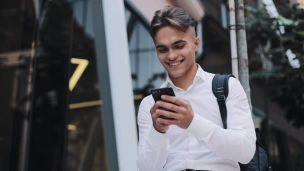 Счастливый молодой бизнесмен или турист с рюкзаком, сидящий рядом с современным офисным зданием по телефону. Коммуникация Успешный богатый портрет Современный красивый смартфон амбициозный — стоковое видео