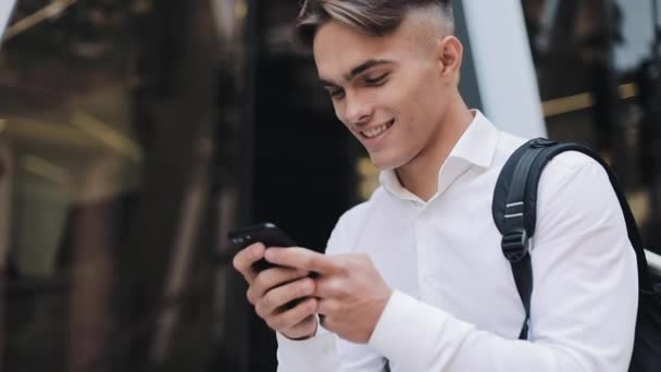 Šťastný mladý podnikatel nebo Turistický batoh pomocí telefonu sedící poblíž moderní kancelářskou budovu. Komunikace úspěšná bohaté portrét moderní pohledný Smartphone ambiciózní — Stock video