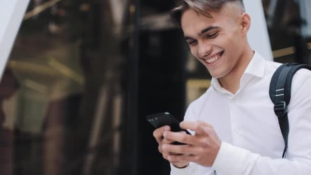 Feliz joven hombre de negocios o turista con una mochila usando el teléfono sentado cerca del moderno edificio de oficinas. Comunicación exitosa rico retrato moderno guapo Smartphone ambicioso — Vídeos de Stock