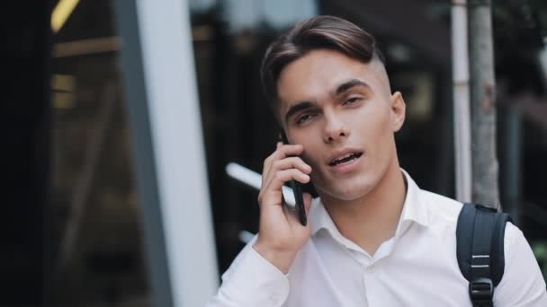 Feliz hombre de negocios ypung con una mochila hablando por teléfono. Se encuentra cerca de un moderno edificio de oficinas. Concepto: tecnología, telefonía, viajes de negocios, negocios — Vídeos de Stock