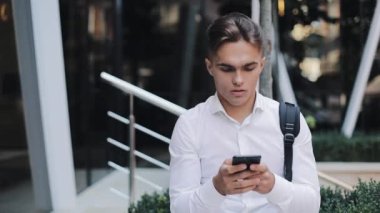 Seriousyoung işadamı veya Turizm Telefon oturma modern Ofis Binası yakınındaki kullanarak bir sırt çantası ile. Konsept: teknoloji, telefon, iş gezileri, iş.