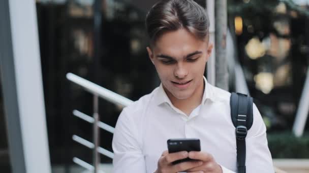 Feliz joven hombre de negocios o turista con una mochila usando el teléfono sentado cerca del moderno edificio de oficinas. Concepto: tecnología, telefonía, viajes de negocios, negocios — Vídeo de stock